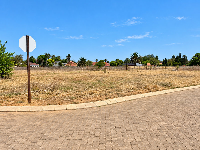 A vacant residential plot featuring a paved street frontage, concrete curbing, and established boundary fencing in a suburban neighborhood setting.