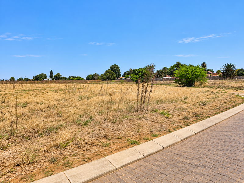 A vacant residential plot featuring dry grass and a paved sidewalk with concrete curbing along the street frontage.