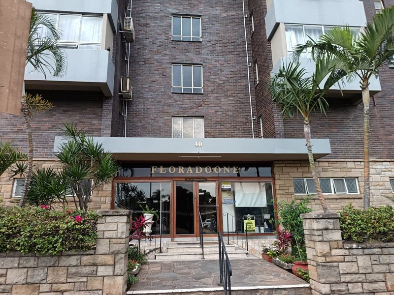 Front entrance view of Floradoone apartment building featuring a brick and stone facade with a glass entryway and paved access path.