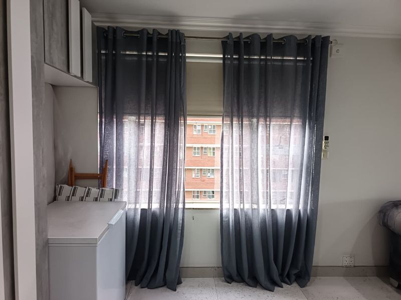 A living area featuring tiled flooring, built-in cabinetry, and a window with curtains overlooking the surrounding residential complex.