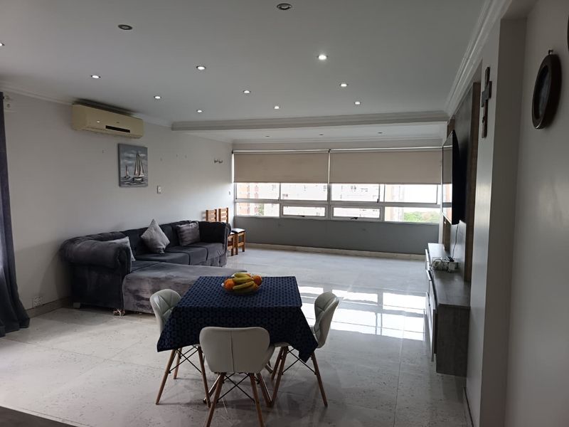 Spacious open-plan living area featuring tiled flooring, recessed downlighting, and a wall-mounted air conditioning unit with large windows providing natural light.