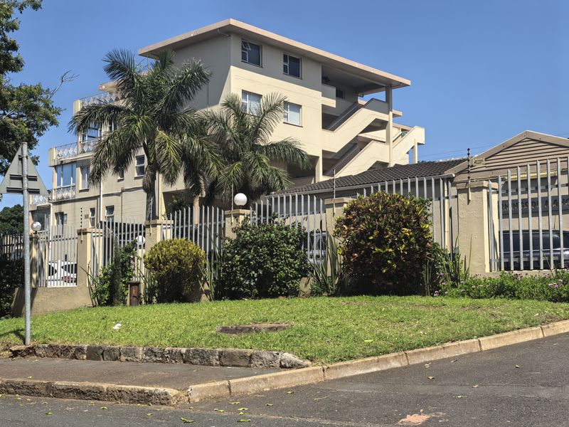 Exterior view of a multi-story apartment building featuring concrete staircases, secure perimeter fencing with electric wires, and landscaped garden area.