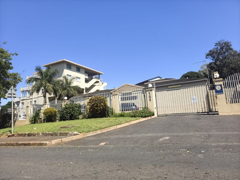 Front exterior view of the apartment complex featuring a secure gated entrance, paved driveway, and well-maintained garden area.
