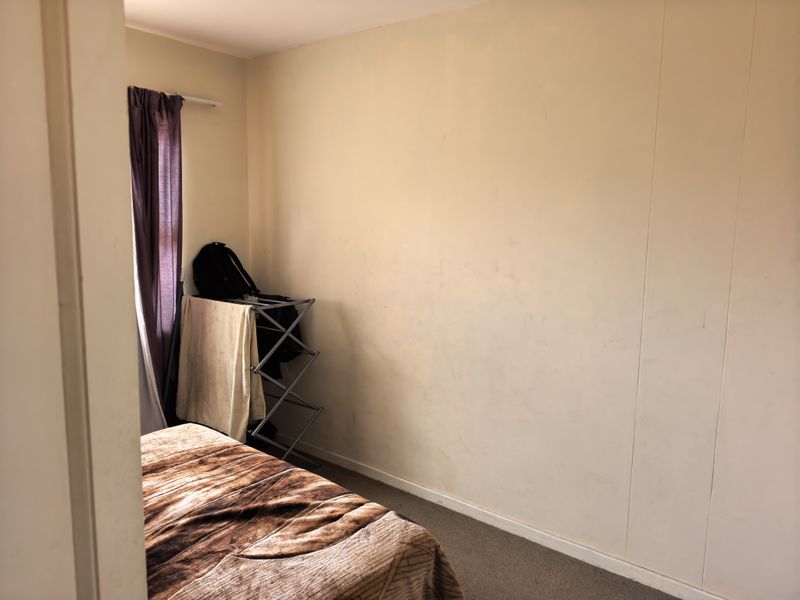 A bedroom featuring carpeted flooring and a window with a curtain rod, currently showing signs of wear on the wall surfaces.