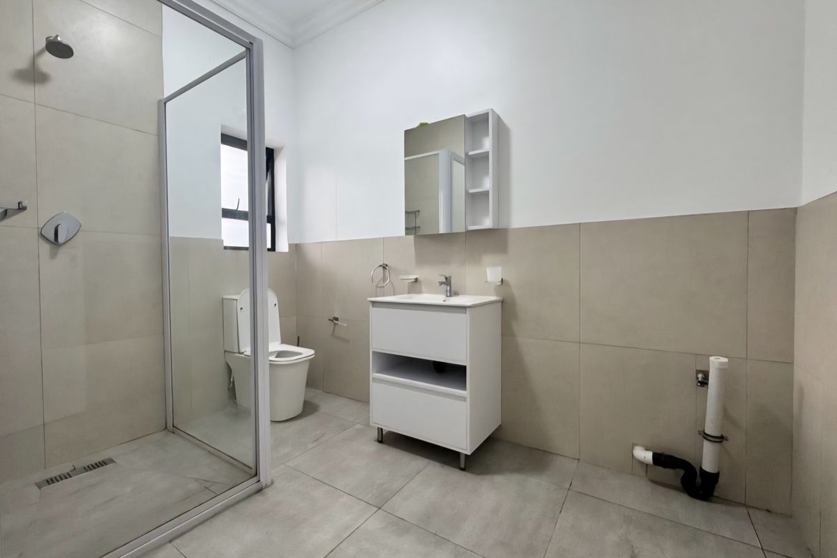 A modern, neutral-toned bathroom featuring a glass-enclosed shower, vanity cabinet with mirror, and toilet. Note the exposed plumbing requiring finishing.