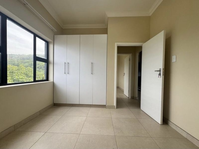 A bright, empty bedroom featuring neutral tiled flooring, built-in white cupboards, and large windows offering natural light and scenic views.