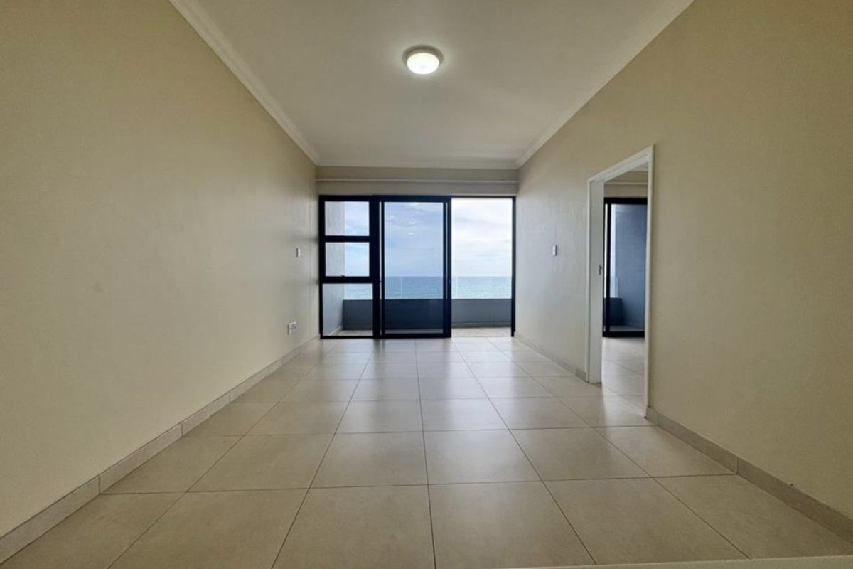 An empty, open-plan living area featuring neutral tiled flooring, cove cornicing, and a large sliding glass door providing access to a balcony with ocean views.