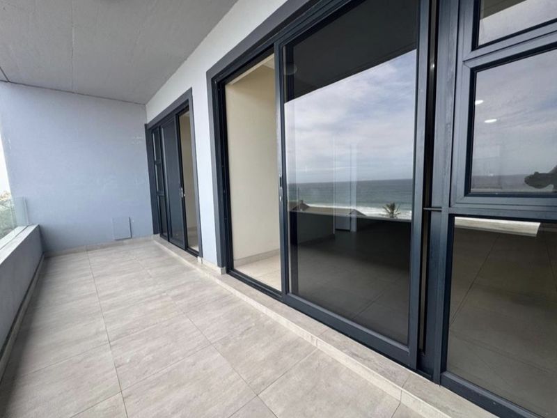A spacious, modern balcony featuring large sliding glass doors, neutral floor tiles, and a glass balustrade with scenic ocean views.