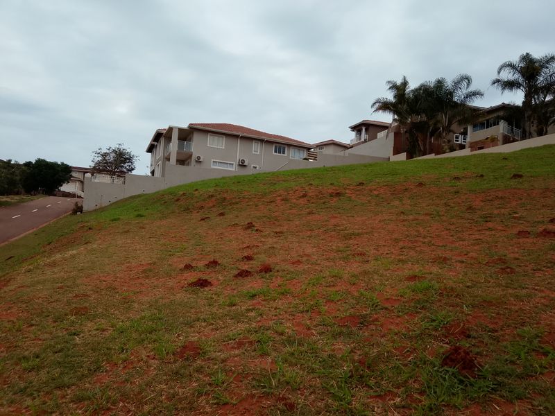 A sloped residential vacant land plot featuring red soil, grass cover, and adjacent paved road access in a developed estate setting.
