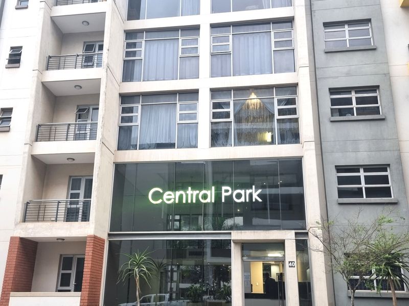 Front elevation of Central Park apartment building featuring a modern glass facade, multiple balconies with metal railings, and well-maintained landscaped garden beds.