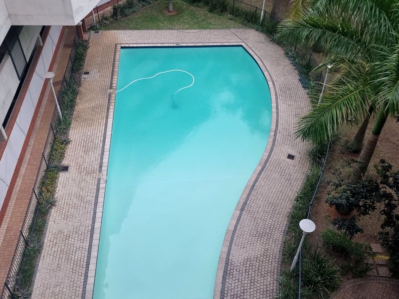 An elevated view of a well-maintained swimming pool area featuring paved decking, perimeter fencing, and established garden landscaping.