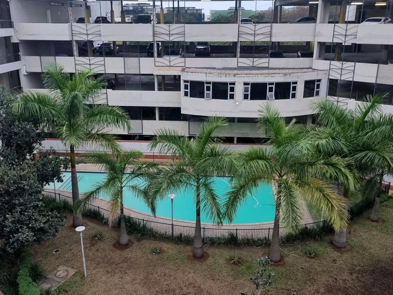An elevated view of a communal swimming pool area set within a landscaped garden, adjacent to a multi-level apartment parking structure.