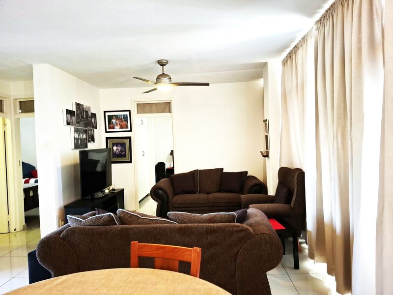 A living room area featuring tiled flooring and a ceiling fan, with natural light entering through large window curtains.