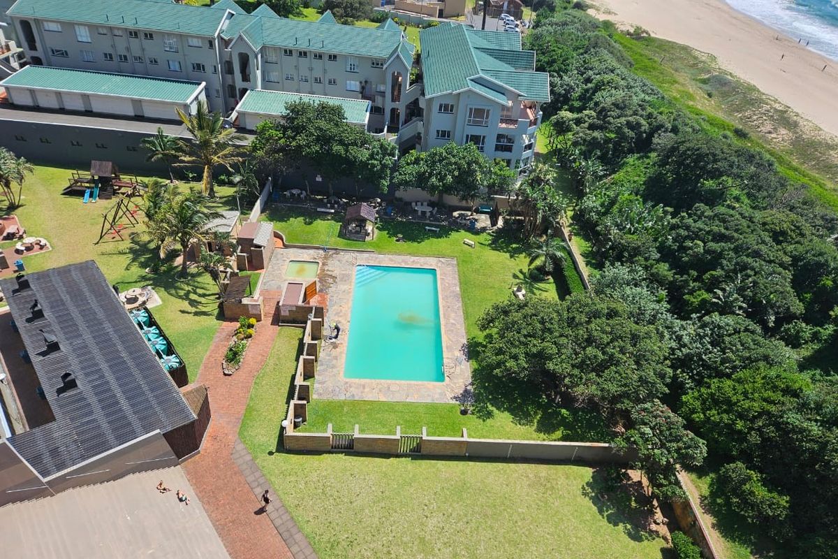 Aerial view of the apartment complex grounds featuring a swimming pool, landscaped garden areas, and paved pathways with direct beach access.
