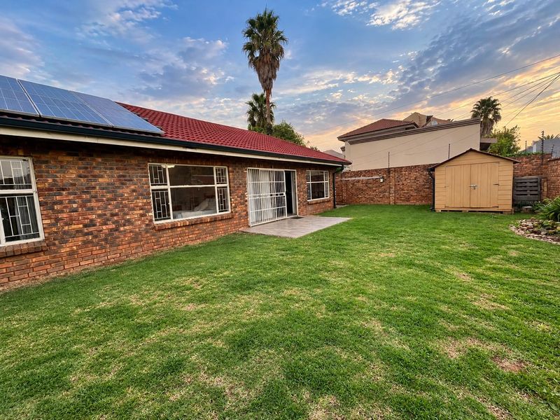Rear view of the property featuring a brick exterior, solar panels on the roof, a well-maintained lawn, and a secure garden shed.