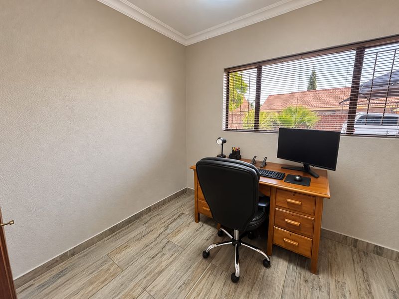 A functional home office featuring wood-look tile flooring, neutral wall finishes, and a large window with horizontal blinds providing natural light.