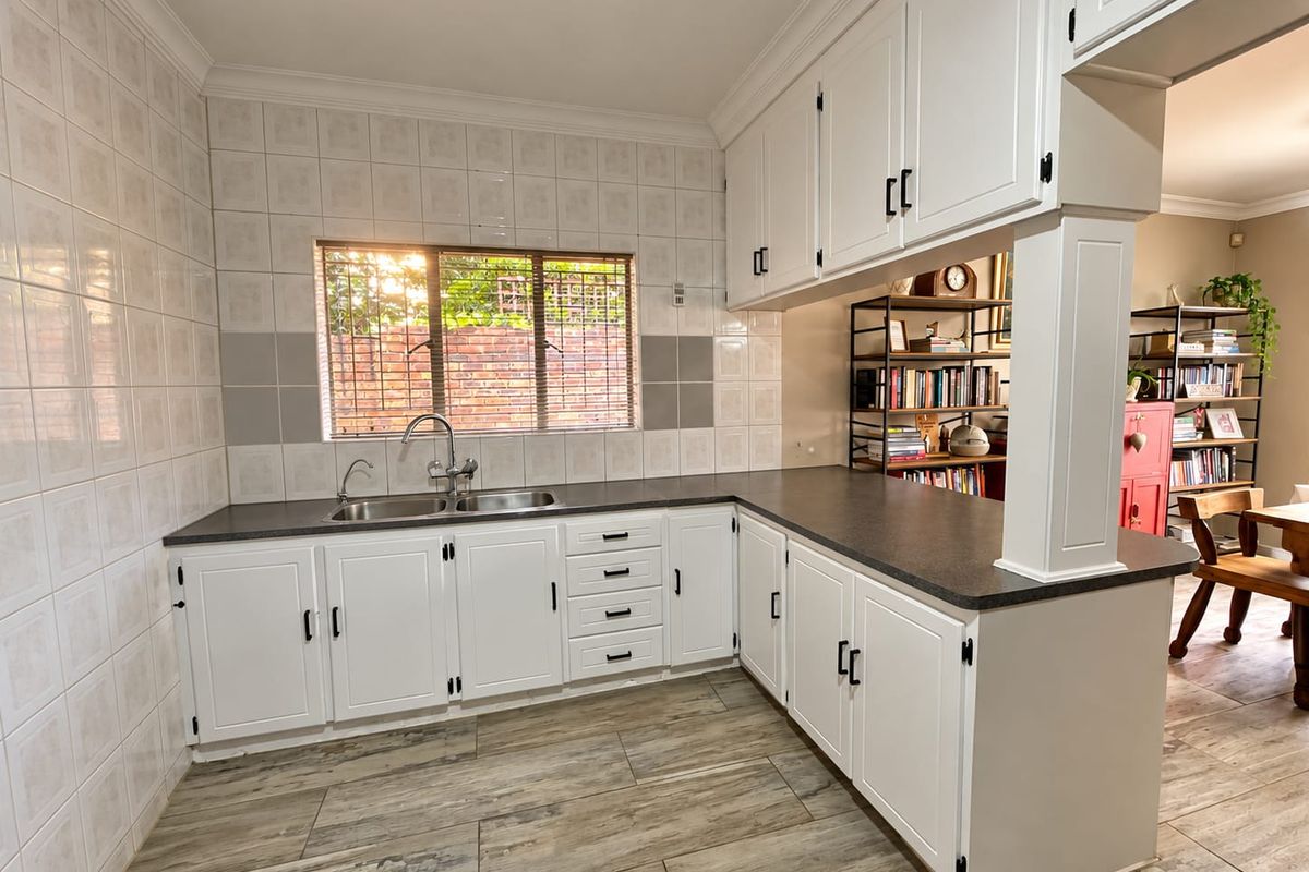 A clean, functional kitchen featuring white cabinetry, dark laminate countertops, a double basin sink, and wood-look floor tiles with an open-plan breakfast bar.