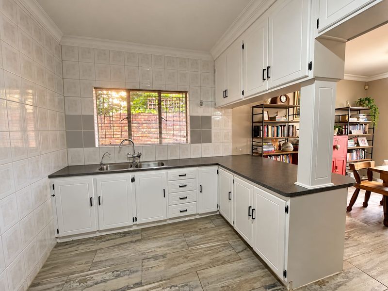 A clean, functional kitchen featuring white cabinetry, dark laminate countertops, a double basin sink, and wood-look floor tiles with an open-plan breakfast bar.