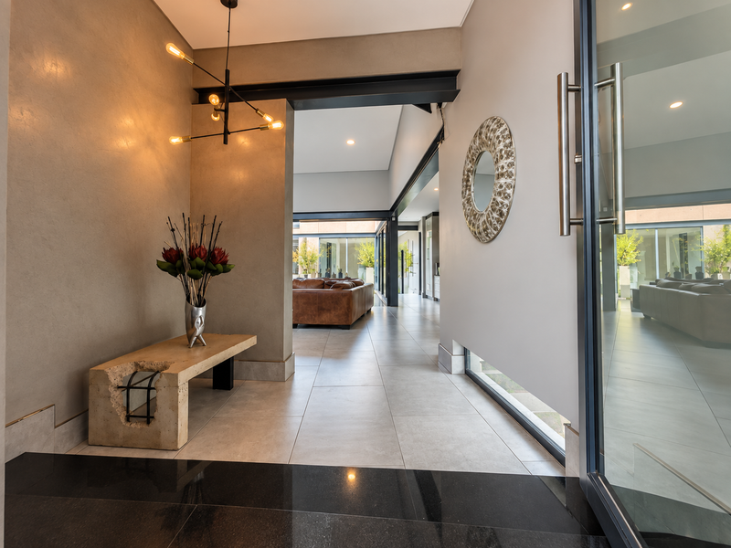 A modern, spacious entrance hall featuring high-quality tiled flooring, an exposed steel beam, and a striking glass pivot door leading to an open-plan living area.