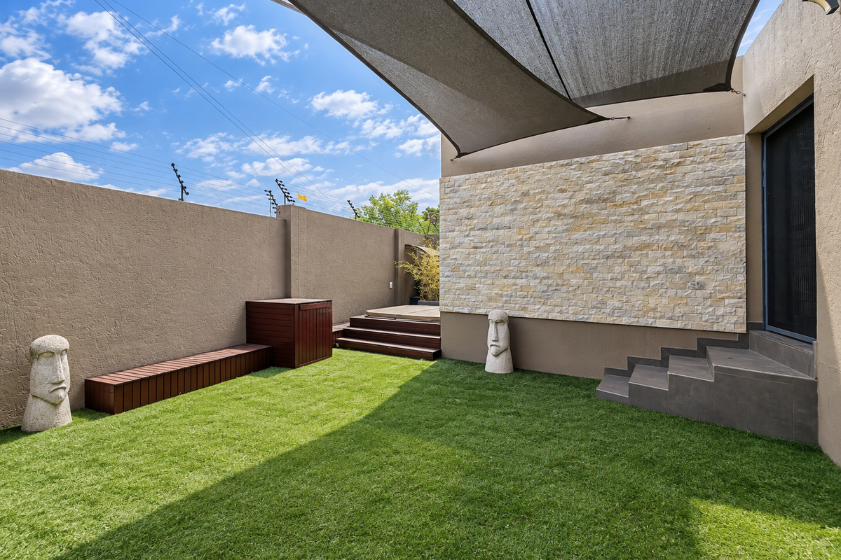 A well-maintained rear garden featuring artificial lawn, timber decking, a stone-clad feature wall, and a large overhead shade sail.