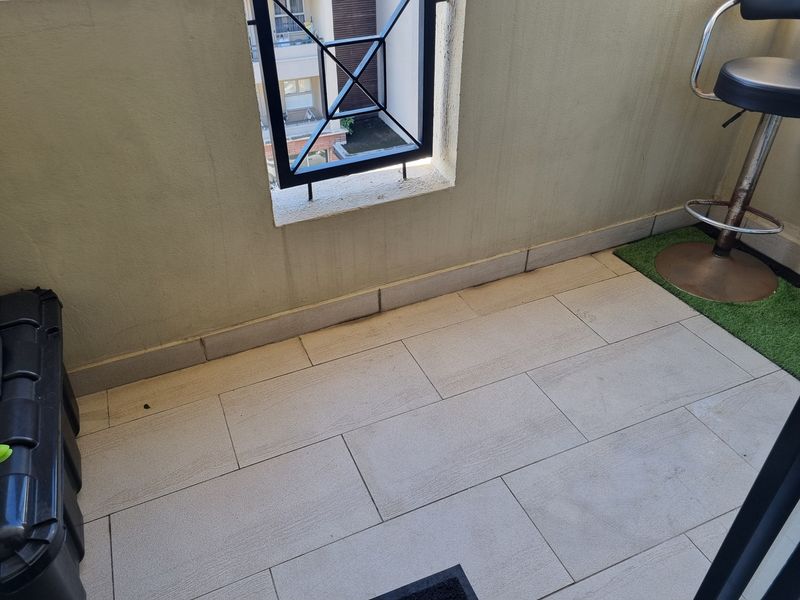 A compact balcony area featuring tiled flooring and a metal security grille, showing some signs of wear on the lower wall sections.
