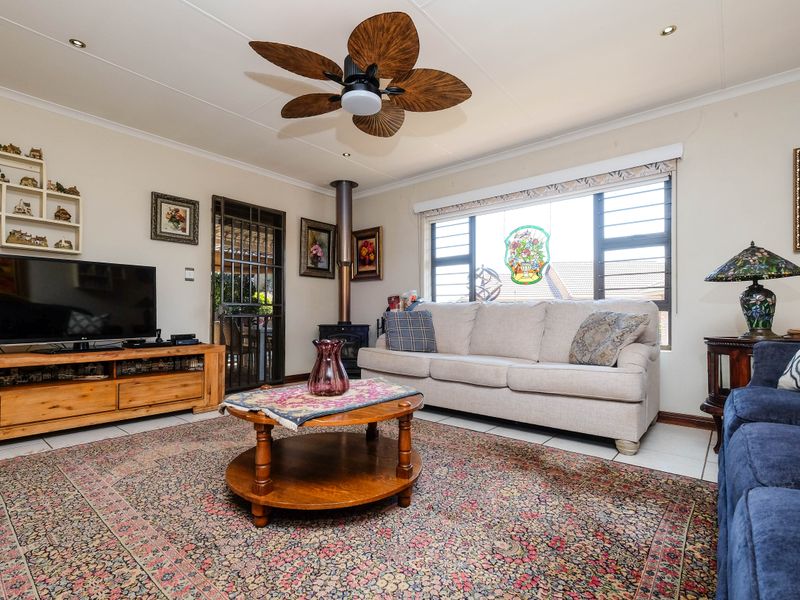A comfortable living room featuring a wood-burning stove, ceiling fan, tiled flooring, and large windows providing natural light.