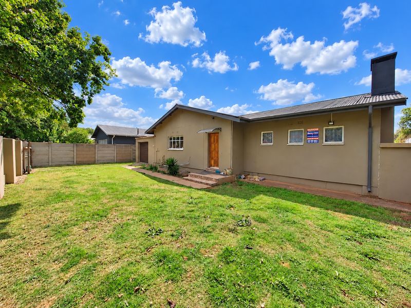 Rear view of the property featuring a spacious lawn, concrete boundary wall, and a single-story structure with a corrugated metal roof and chimney.