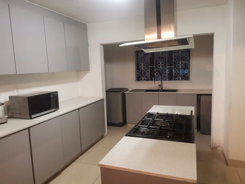 Modern kitchen featuring grey cabinetry, stone countertops, a central gas hob with extractor fan, and a separate scullery area with a double sink.