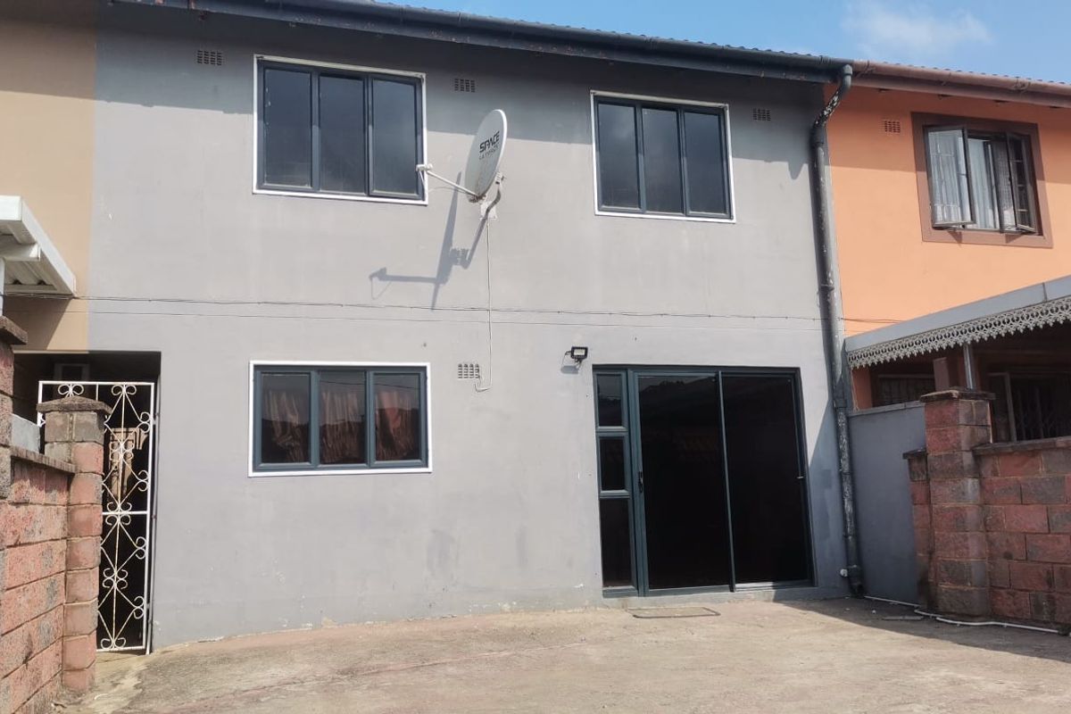 Rear exterior view of a two-story residential property featuring grey painted walls, dark aluminium window frames, a sliding glass door, and paved courtyard area.