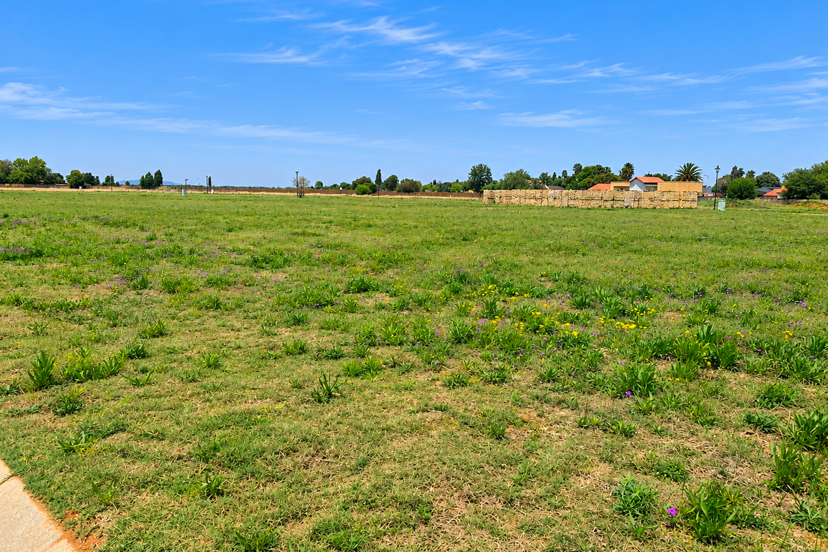 A wide, open residential vacant land plot featuring natural grass coverage and a clear boundary wall in the distance.