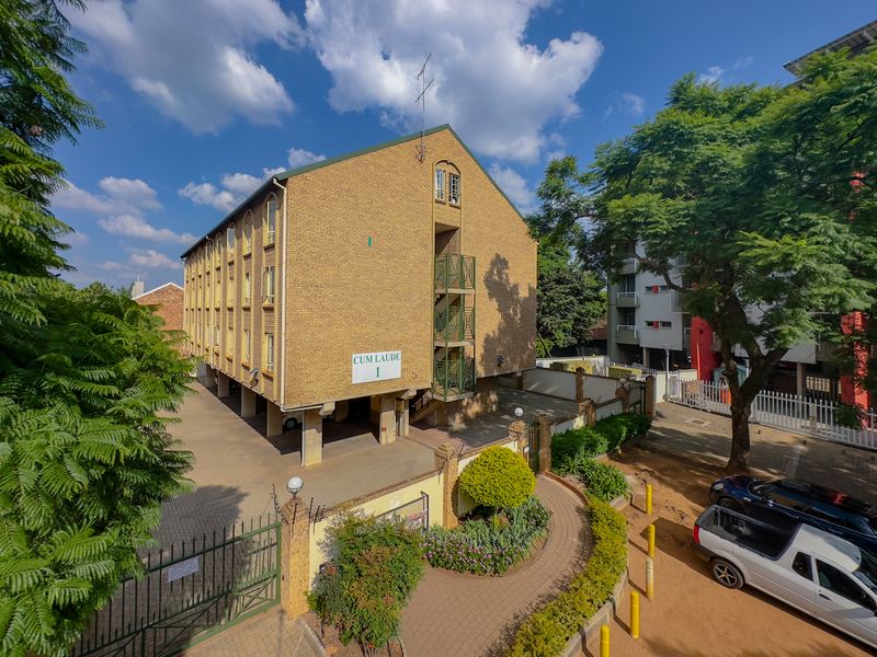Exterior view of a multi-story face brick apartment building featuring a stilt-supported ground level, metal staircases, and a landscaped front garden area.