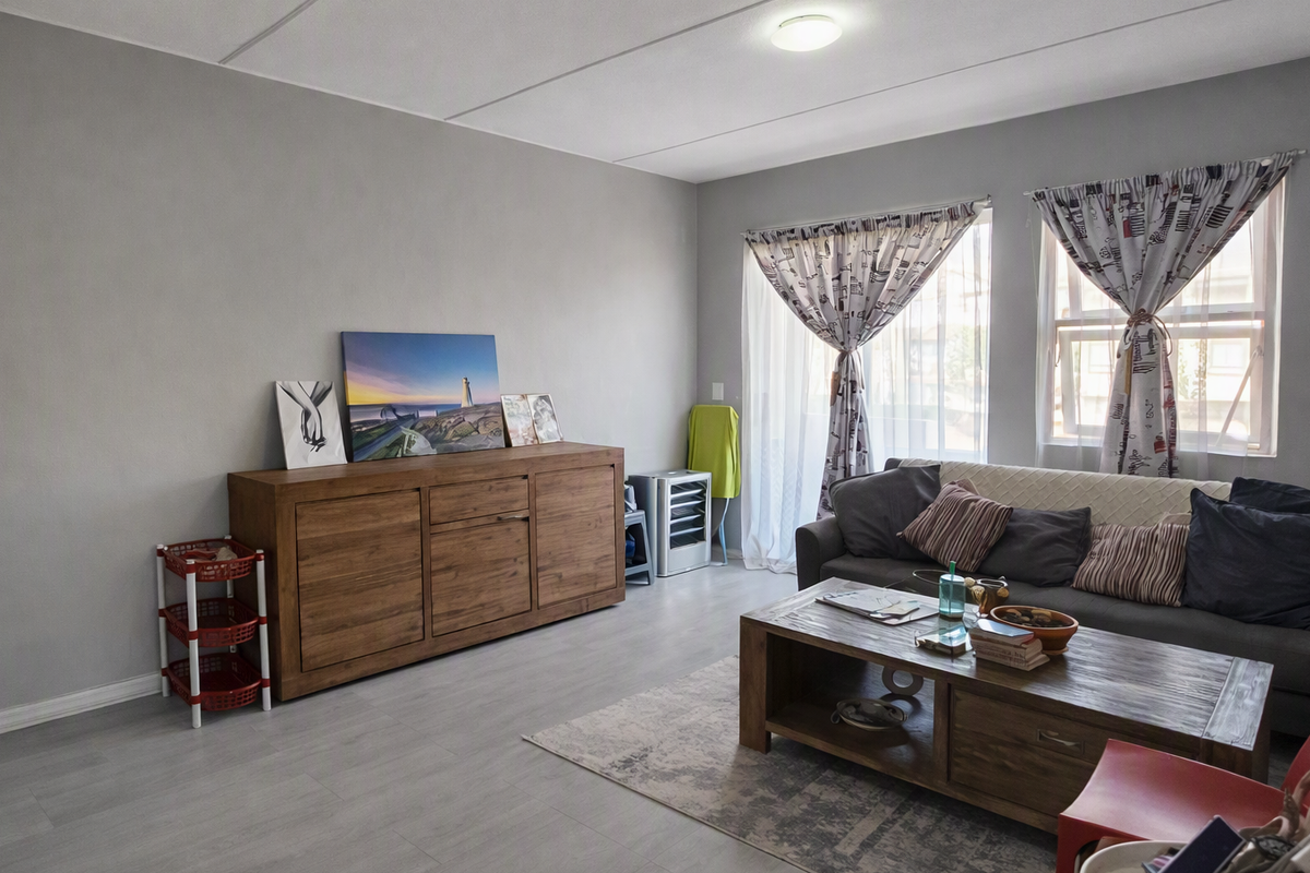 A well-maintained living room featuring light grey laminate flooring, neutral wall tones, and natural light provided by a sliding door and dual windows.