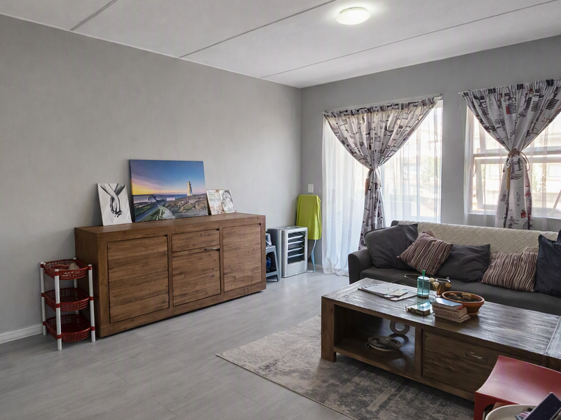 A well-maintained living room featuring light grey laminate flooring, neutral wall tones, and natural light provided by a sliding door and dual windows.