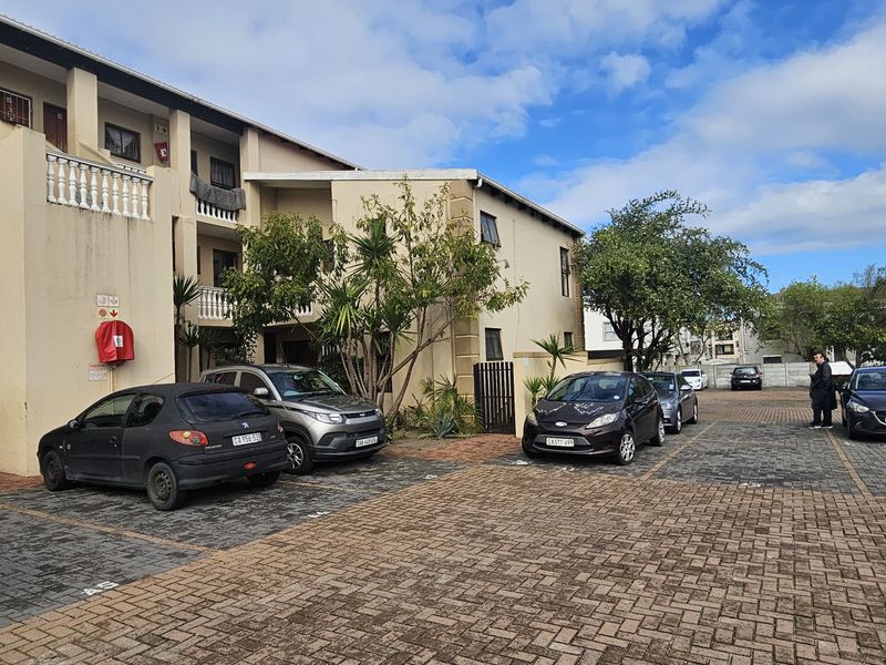 Exterior view of an apartment complex featuring a paved parking area, multi-level balconies with balustrades, and established landscaping.