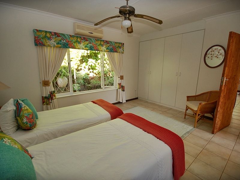 A bedroom featuring built-in cupboards, tiled flooring, a ceiling fan, and an air conditioning unit, with a window overlooking the garden.