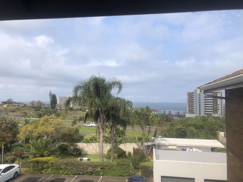 An elevated view overlooking a suburban landscape with greenery, nearby buildings, and a distant ocean horizon.