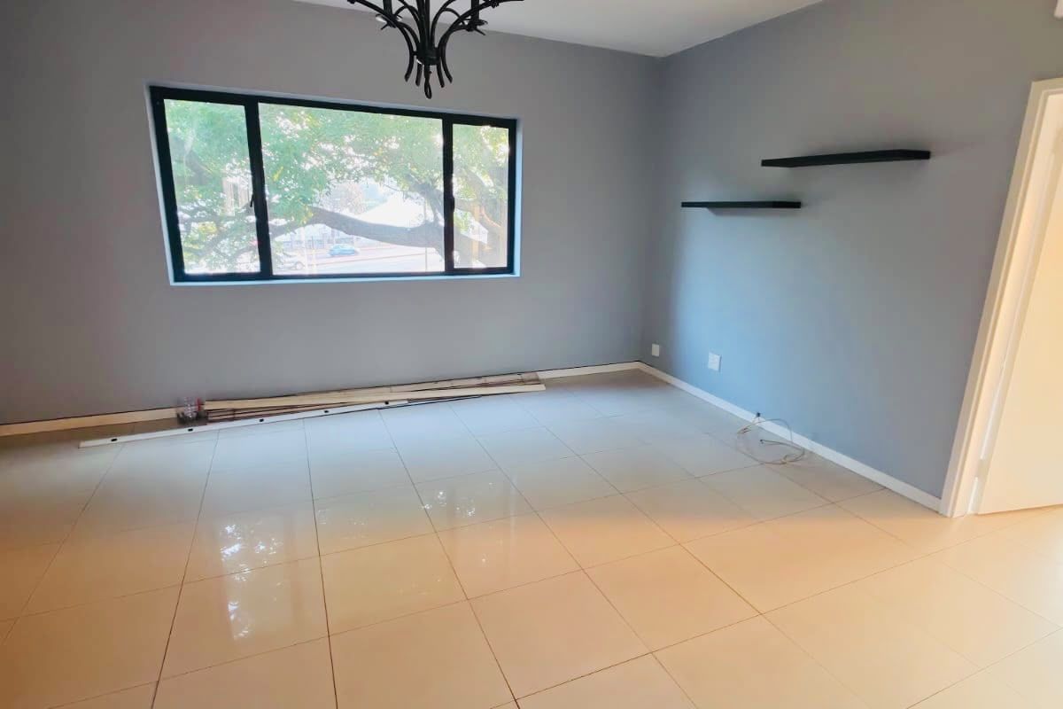 A bright, empty living room featuring neutral grey walls, polished porcelain floor tiles, a decorative chandelier, and large black-framed windows.