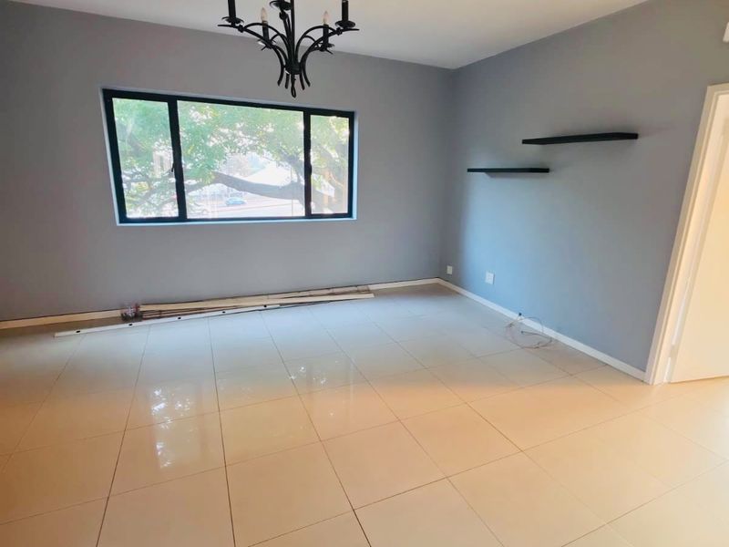 A bright, empty living room featuring neutral grey walls, polished porcelain floor tiles, a decorative chandelier, and large black-framed windows.