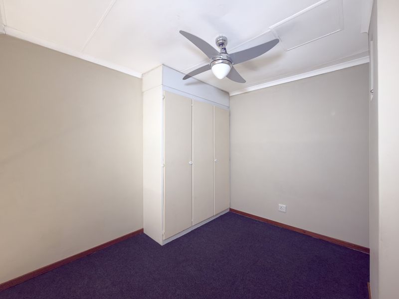 A clean, empty bedroom featuring neutral walls, dark carpeting, a built-in wardrobe, and a ceiling fan.