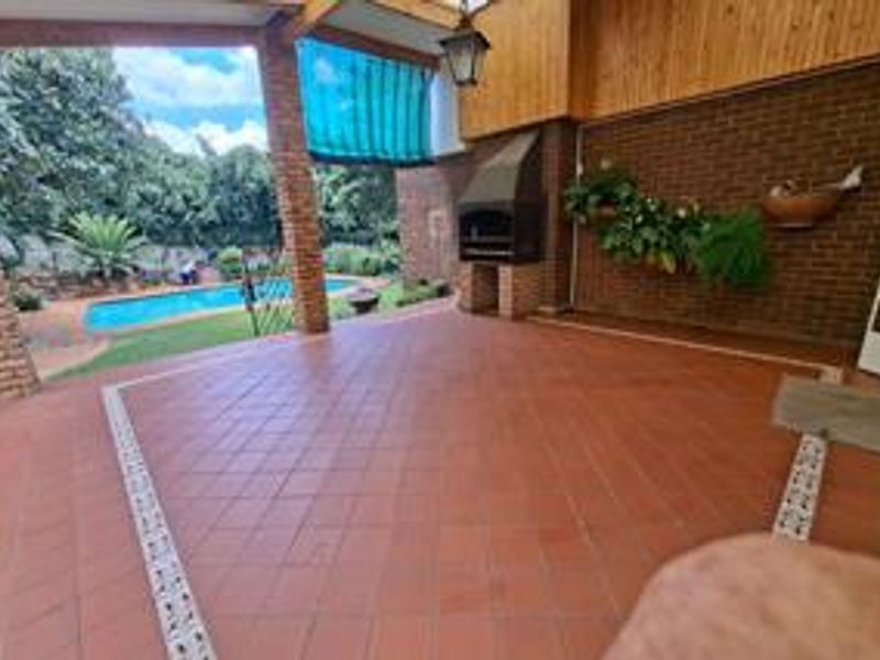 Covered outdoor patio featuring a built-in braai, brick pillars, and tiled flooring, overlooking the swimming pool and garden area.