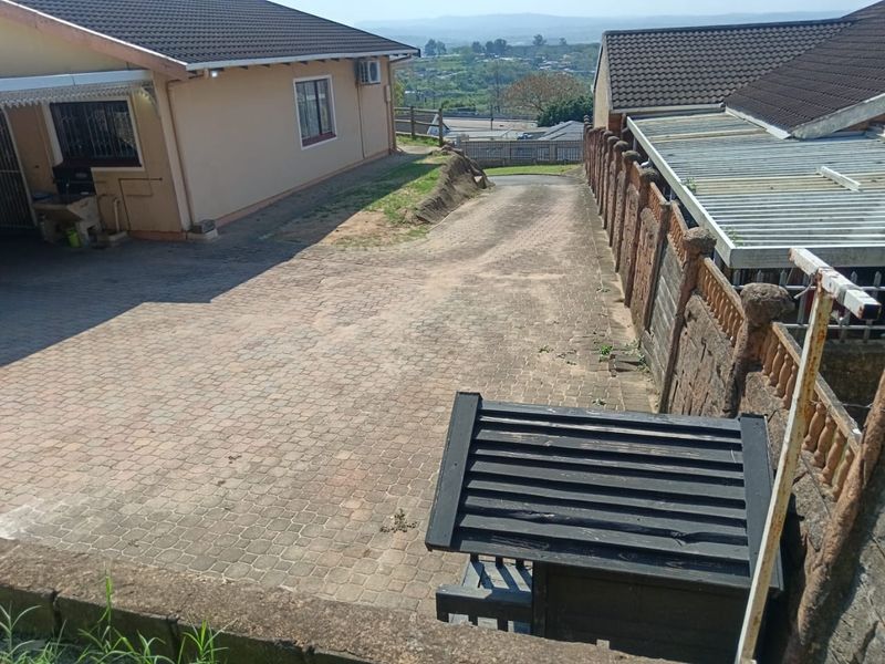 A view of the paved driveway and front exterior of the house, featuring a tiled roof and perimeter walling with distant landscape views.