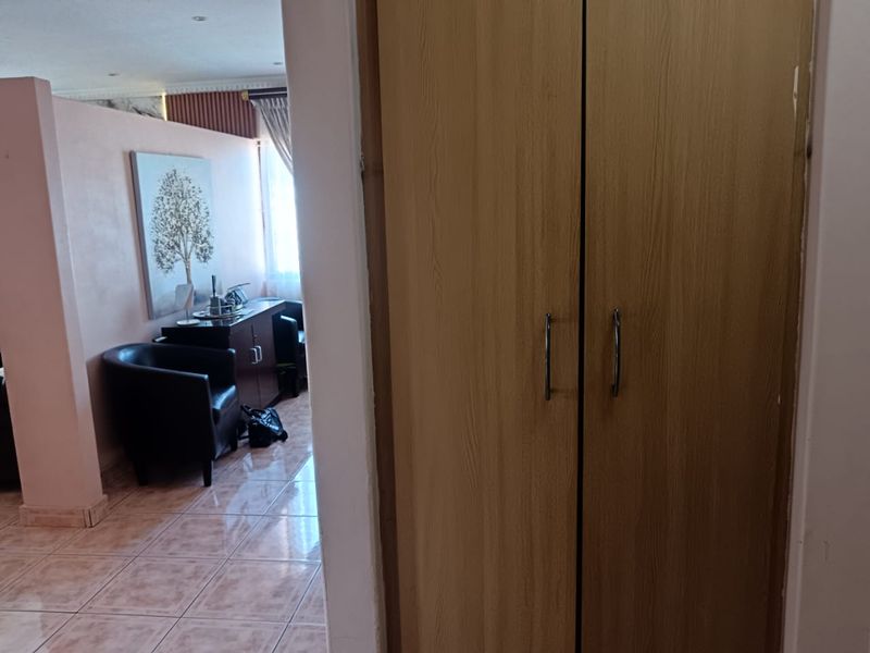 A bedroom interior featuring a built-in wooden wardrobe and tiled flooring, with a view into an adjacent living area.