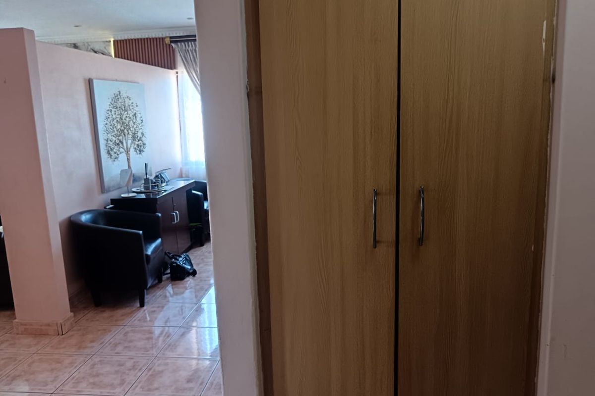 A bedroom interior featuring a built-in wooden wardrobe and tiled flooring, with a view into an adjacent living area.