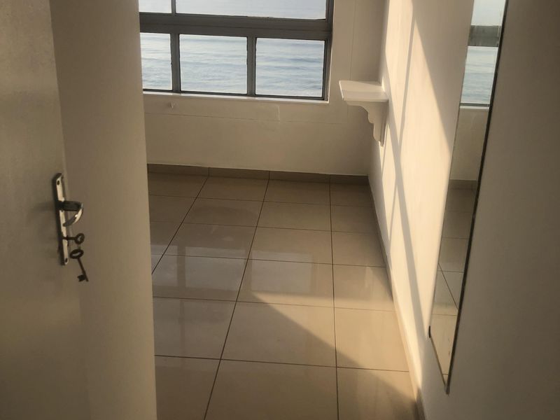 A bright, empty bedroom featuring tiled flooring, a wall-mounted shelf, and a large window offering scenic ocean views.