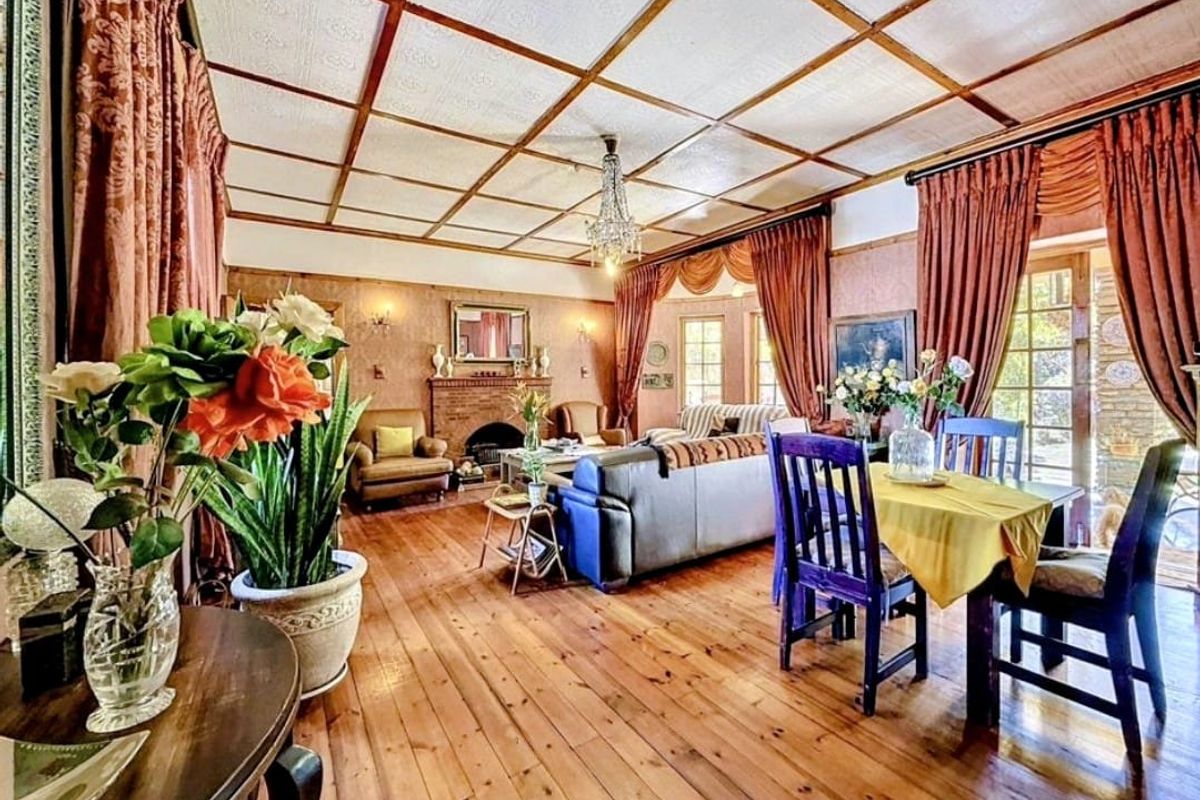 Spacious open-plan living and dining area featuring warm timber flooring, a classic brick fireplace, and a distinctive coffered ceiling with a central chandelier.