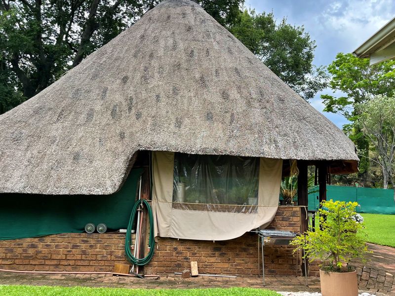 A rear view of a rustic structure featuring a traditional thatch roof and face brick walls, set within a garden area.