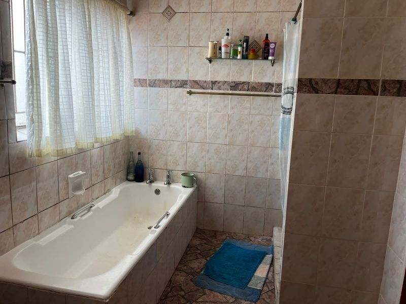 A dated bathroom featuring a built-in bathtub with shower attachment, tiled walls and flooring, and a glass storage shelf.