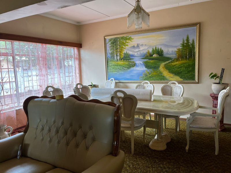 A dining room featuring a large window with security bars and a central ceiling light fixture, currently showing dated finishes and carpeting.