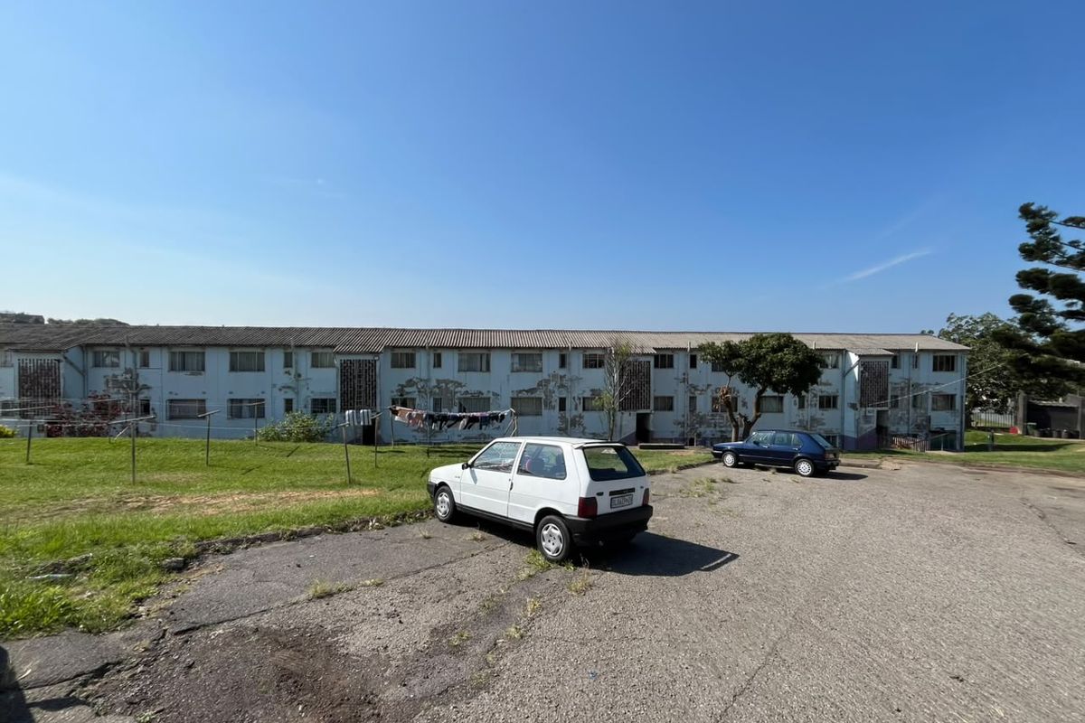 Exterior view of a multi-story apartment block featuring a communal lawn and asphalt parking area, showing signs of significant exterior wear.