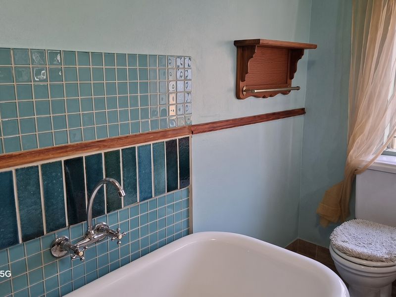 A bathroom featuring a wall-mounted faucet, decorative tiled splashback, built-in bathtub, and a standard toilet with window security bars.